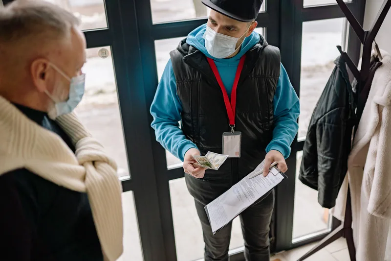 A masked delivery courier receiving cash payment from a customer at the doorstep
