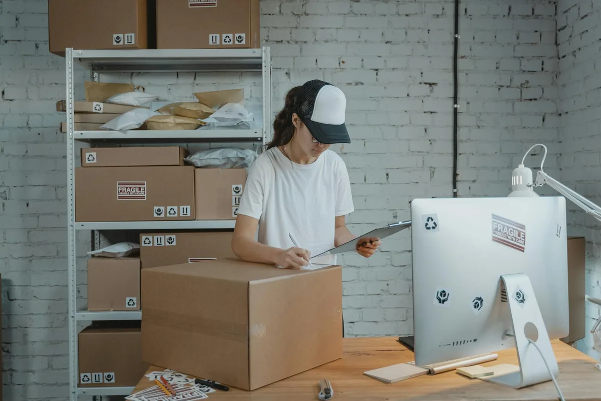Malaysian ecommerce seller at a packing station comparing Ninja Van and J&T Express courier options on a laptop with shipping boxes in the background