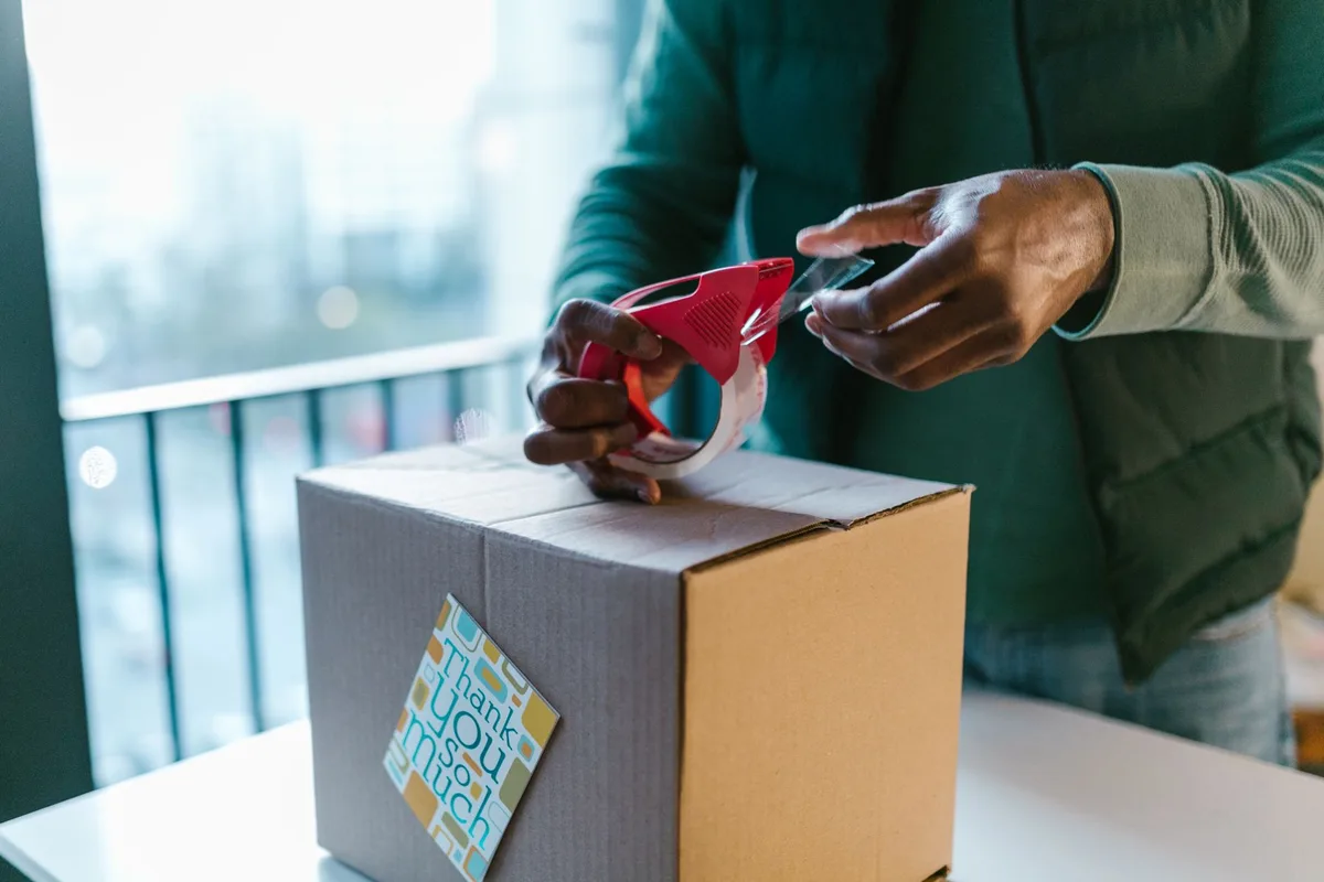 An ecommerce seller sealing a cardboard shipping box with a tape gun before calculating the shipping cost
